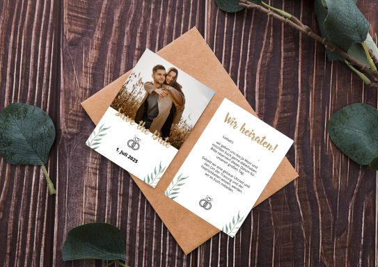 Save the Date Karte Zwei Karten liegen auf einem braunen Umschlag, der sich auf einem Holztisch mit Eukalyptusblättern befindet.